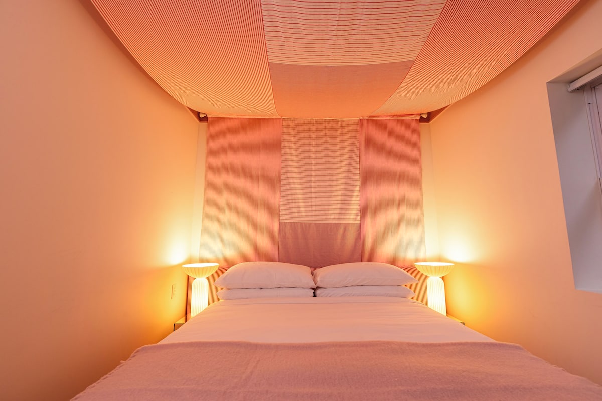 Cama bem arrumada com lençóis brancos, duas luminárias de cabeceira e cortina listrada.