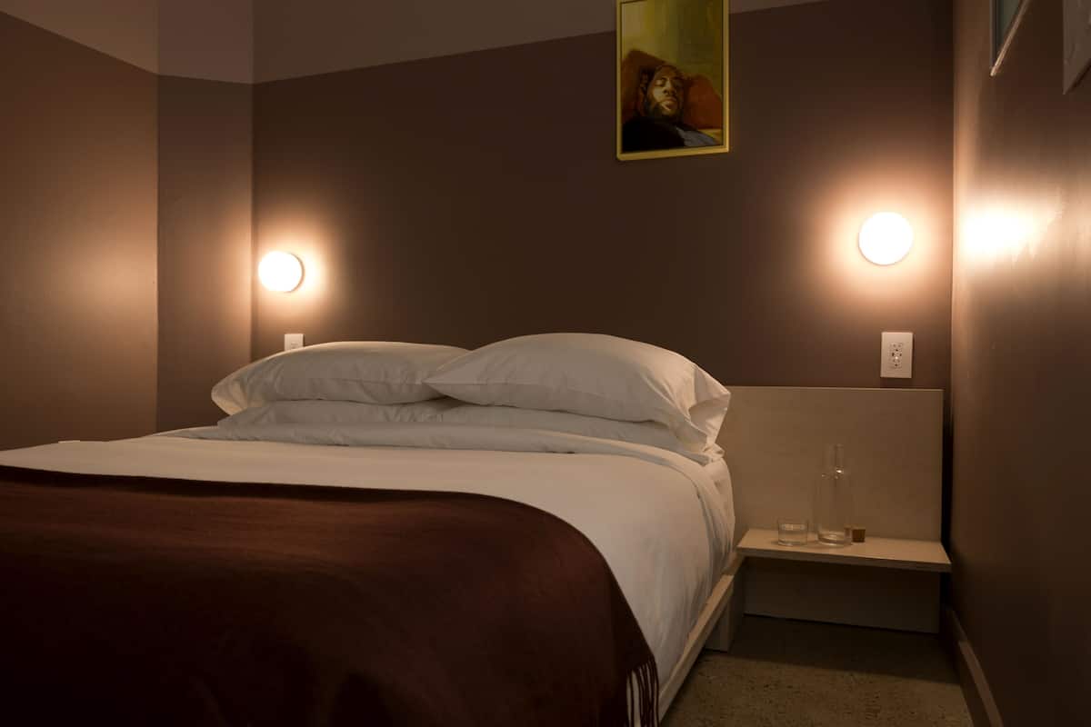 Uma cama bem arrumada com lençóis brancos e um cobertor marrom, uma mesa de cabeceira com um copo e uma garrafa, e duas luminárias fixadas na parede.
