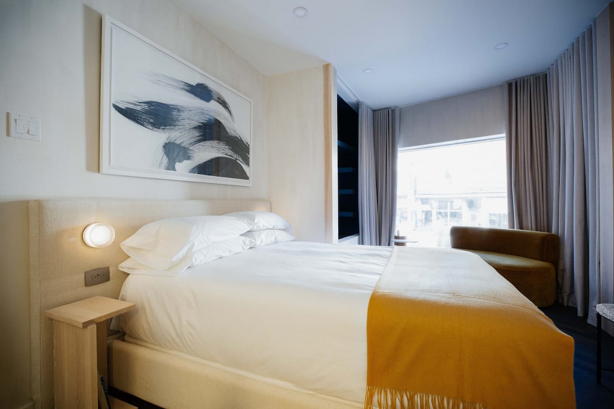 Quarto de hotel com cama, mesa de cabeceira, cadeira, janela grande e um quadro na parede.