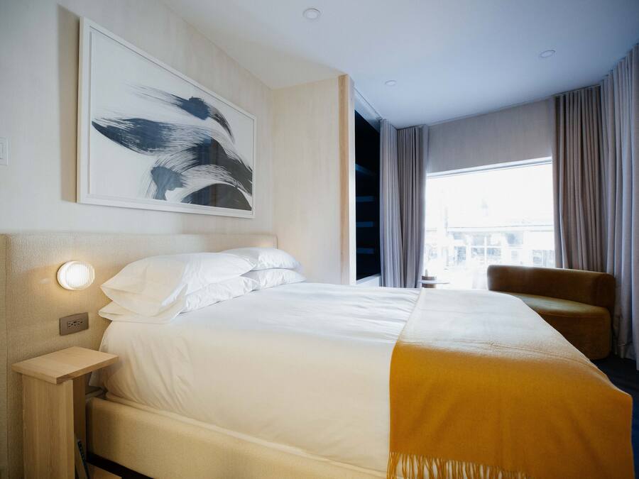 A hotel room with a bed, a bedside table, a chair, a large window, and a painting on the wall.