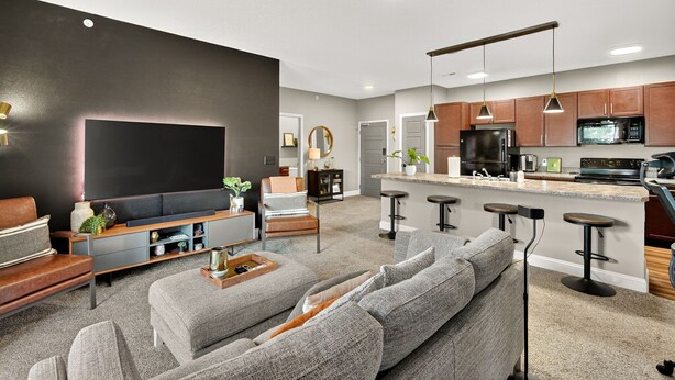 A modern living room with a large flat-screen TV, a grey sectional sofa, and a coffee table. The kitchen features dark wooden cabinets, a black refrigerator, and a microwave.