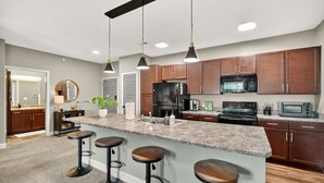 A modern kitchen with granite countertops, dark wooden cabinets, a stainless steel refrigerator, a microwave, and a built-in oven. There are three bar stools at the island.