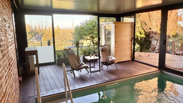 Zona de piscina interior moderna amb terrassa de fusta, seients relaxants i una porta de vidre que dona a un jardí exterior.