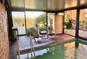 A modern indoor pool area with wooden decking, lounge chairs, and a glass door leading to an outdoor garden. - Entre Terre et Estuaire & Spa (La Riviere-Saint-Sauveur)