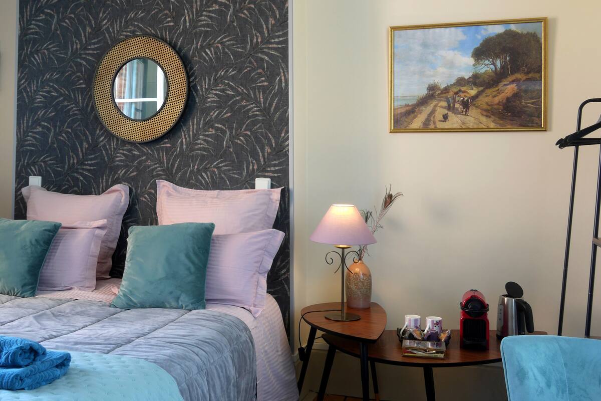 Een slaapkamer met een bed, twee nachtkastjes, een ronde spiegel, een schilderij en een stoel.