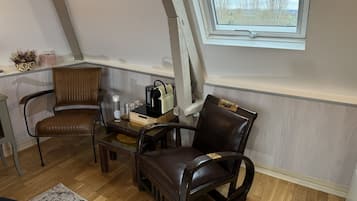 Un acogedor espacio tipo loft con suelo de madera, dos sillones de cuero, una pequeña mesa de madera y un ventanal con vistas a árboles y un cielo nublado.