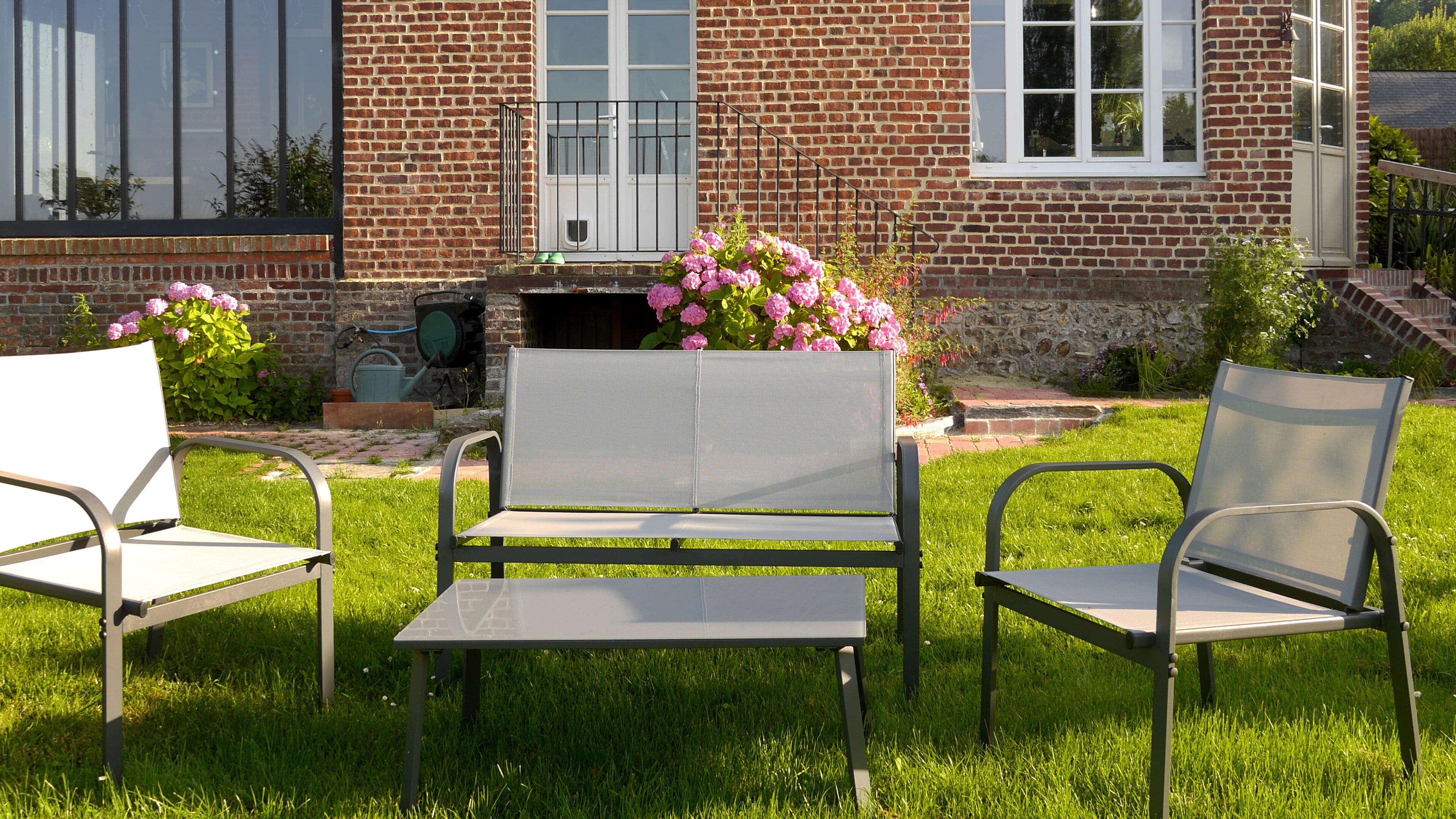 Eine Terrasse mit zwei Metallstühlen und einem Tisch, ein Ziegelhaus mit einem Fenster und ein Garten mit rosa Blumen.