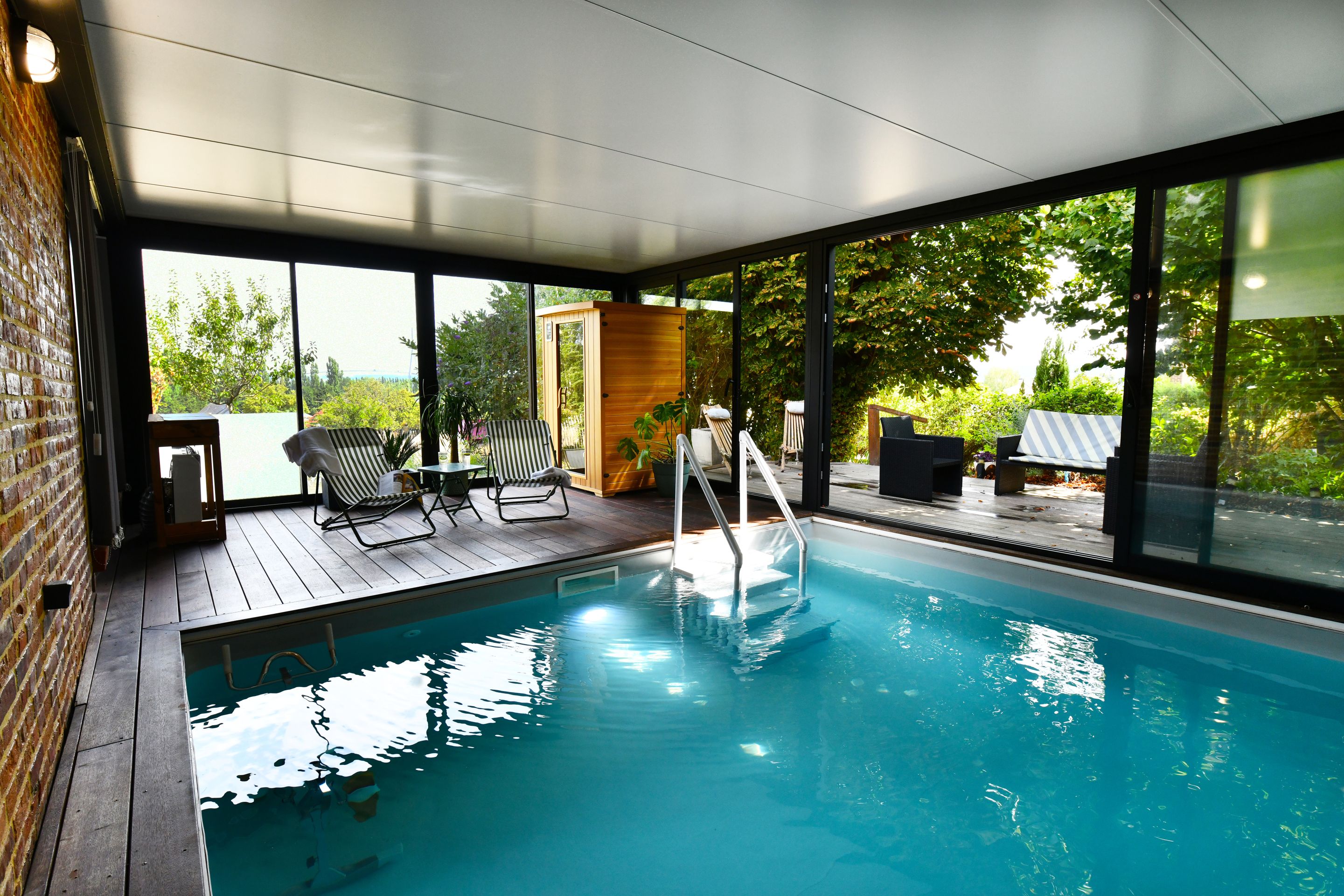 Una piscina interna moderna con pareti in vetro trasparente, un pavimento in legno e poltrone relax.