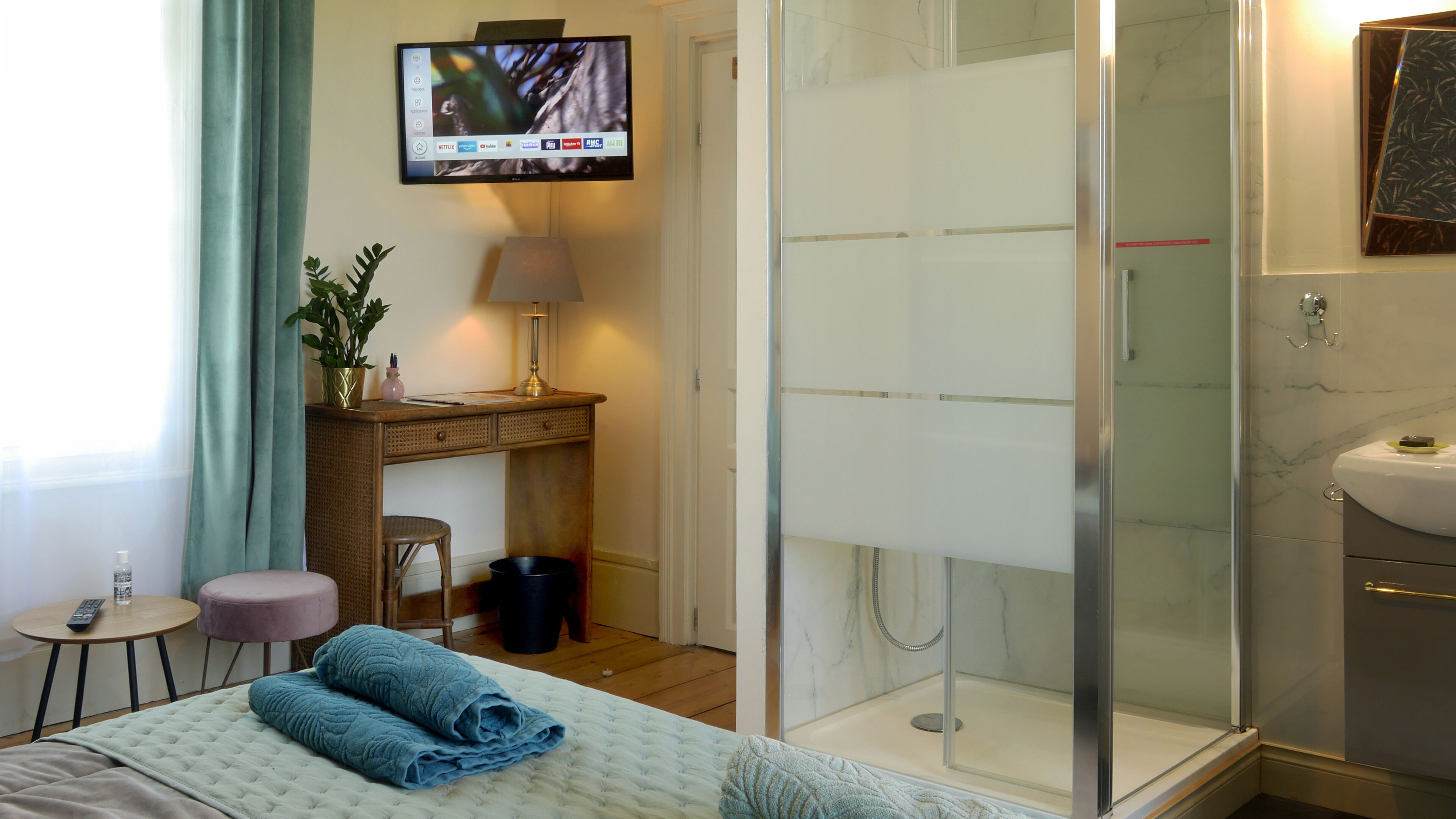 A hotel room with a bed, a desk with a lamp, a TV mounted on the wall, a shower, and a bathroom with a sink.