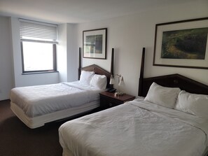 Habitación de hotel con dos camas, una ventana con persianas, una mesita de noche con lámpara y cuadros en las paredes.
