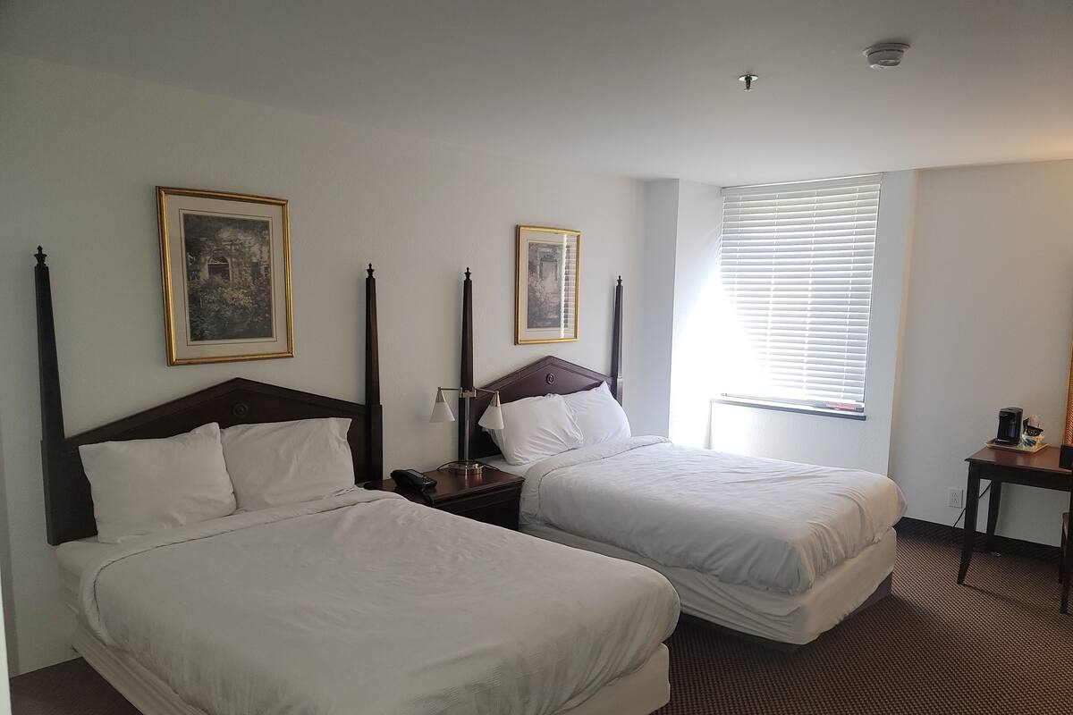 A hotel room with two beds, a nightstand with a lamp, a framed picture on the wall, and a window with blinds.