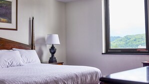 A hotel room with a bed, a nightstand with a lamp, a framed picture on the wall, and a window with a view of greenery. - The McLure Hotel and Conference Center (Wheeling)