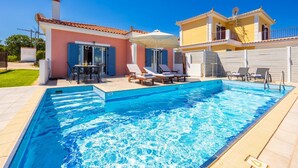 Una piscina con lettini e un tavolo, circondata da una casa con tetto in tegole e un giardino.
