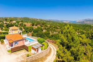 A two-story house with a swimming pool, surrounded by greenery and hills.