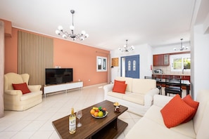 A modern living room with a flat-screen TV, sofas, a coffee table with fruit, and a kitchen in the background. - Villa Europe Ena - Two Bedroom Villa, Sleeps 4 (Ionian Islands)