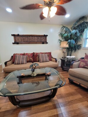 A living room with a glass coffee table, a sofa with patterned cushions, a ceiling fan, and a large potted plant. - The "Tommy Suite". North end of the Island! 350 yrds. from The Gulf of America! (Fort Myers Beach)