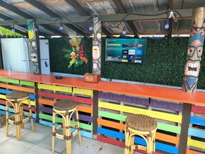 A bar area with a TV screen displaying a menu, colorful striped seating, and tiki decorations.