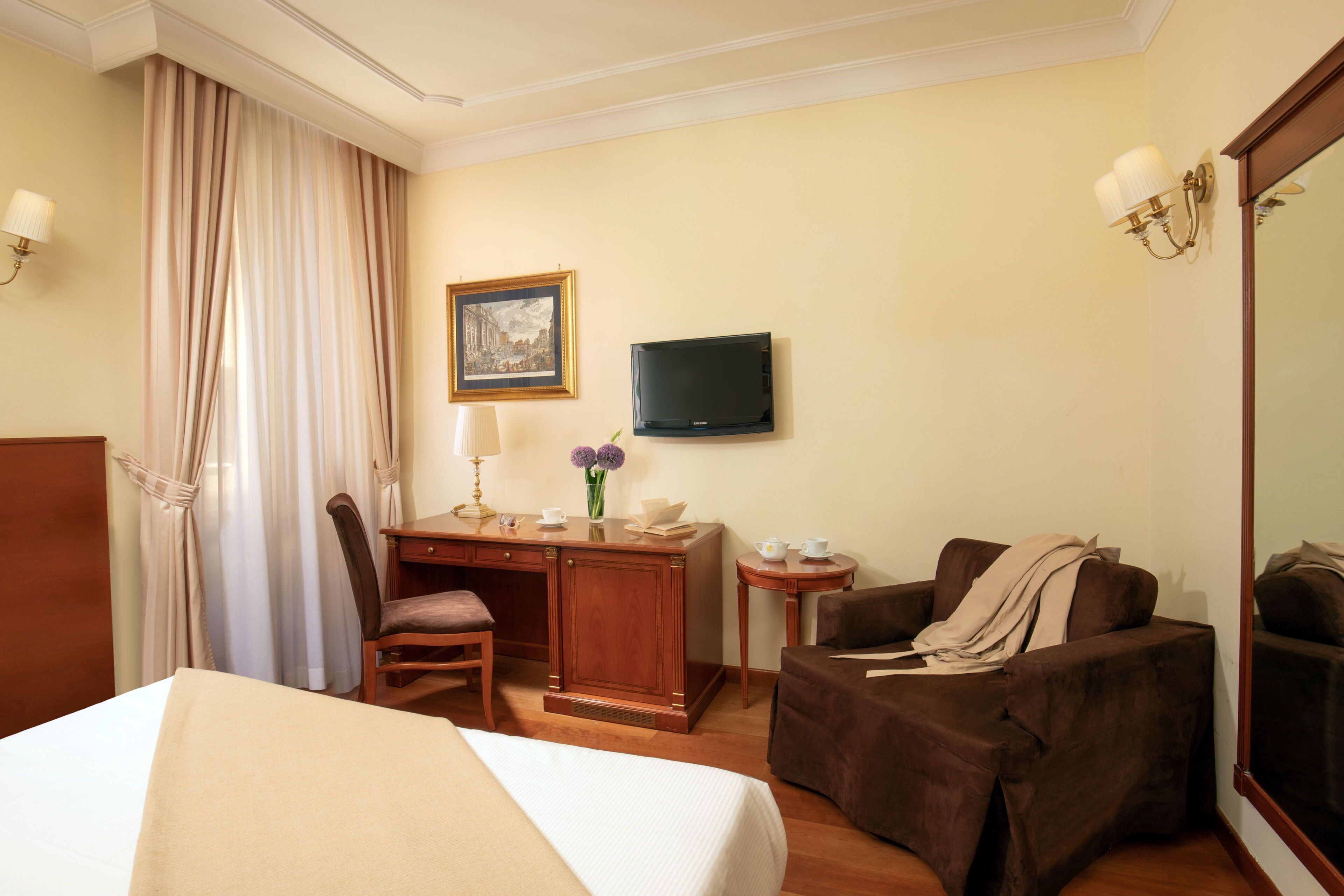 Una habitación de hotel con una cama, un escritorio y una silla, una mesita con lámpara, televisión y un espejo grande.