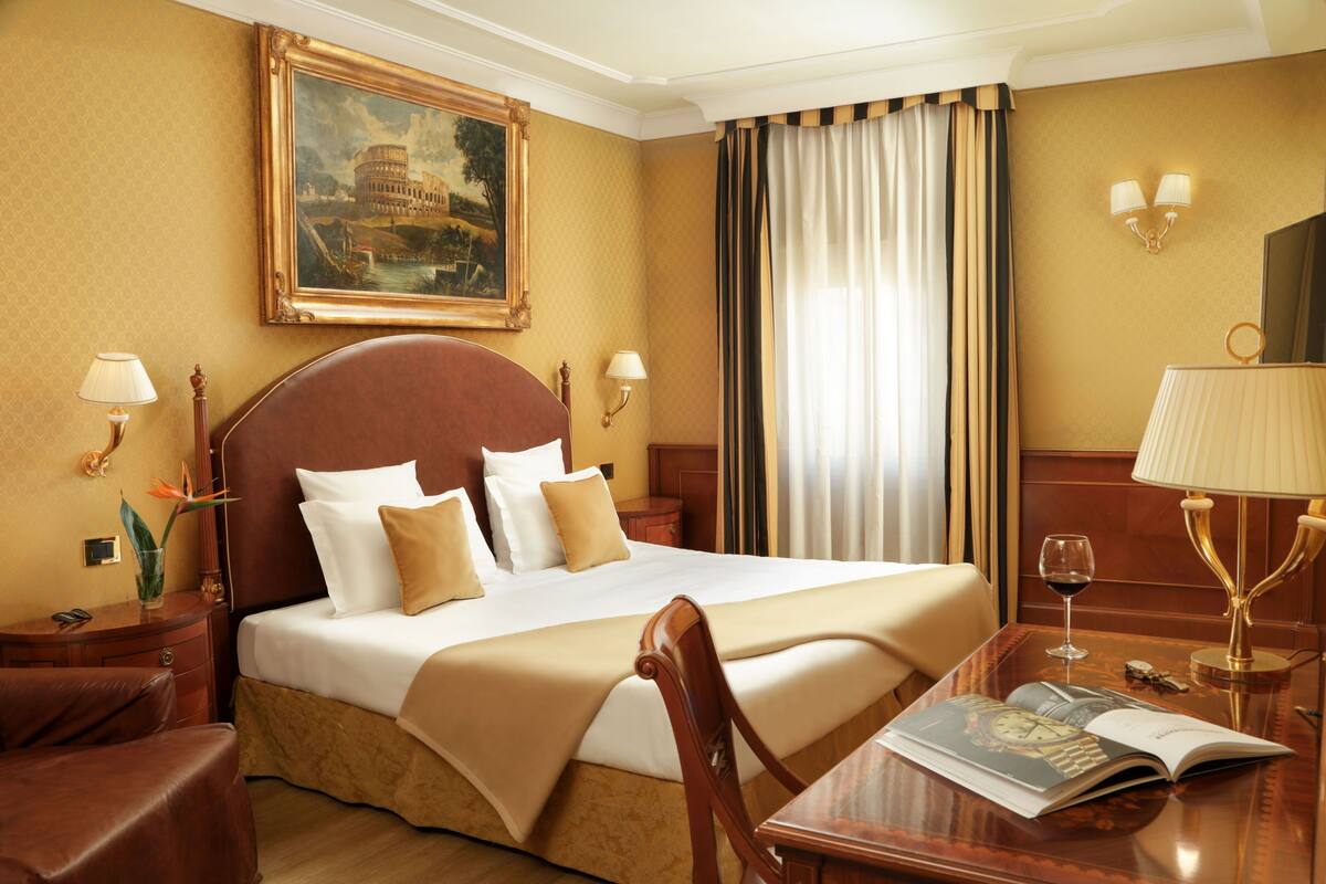 Una camera d'albergo con un letto grande, una testiera in legno, due lampade da comodino, un comodino, una scrivania con una rivista e un quadro del Colosseo appeso al muro.