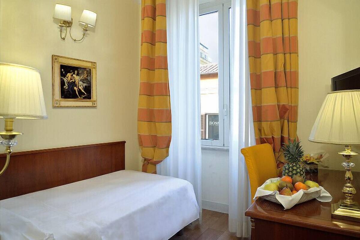 Una camera d'albergo con un letto, due lampade a parete, un quadro incorniciato, una finestra con tende, una sedia e un tavolo con della frutta.