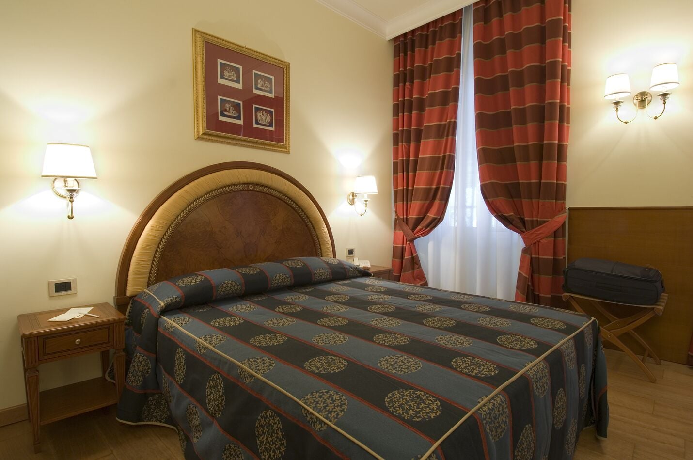 A hotel room with a large bed, a wooden headboard, two bedside lamps, a nightstand, a desk with a chair, and a window with striped curtains.