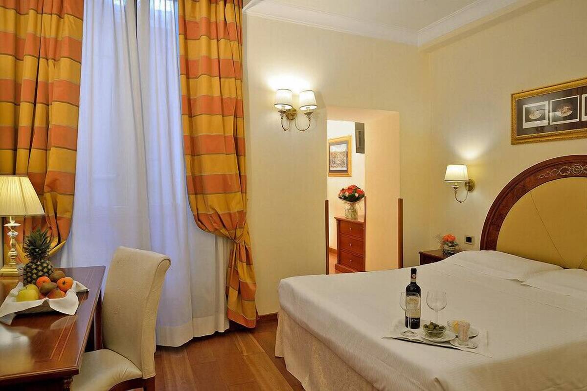 Una camera d'albergo con un letto, una scrivania con una ciotola di frutta, una lampada e una finestra con tende.