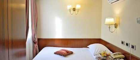 Habitación de hotel con cama, mesita de noche, lámpara, unidad de aire acondicionado y teléfono en la pared.