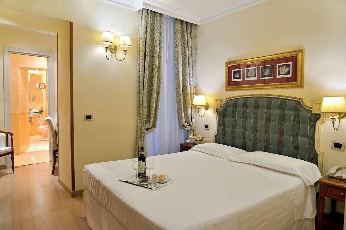 Camera d'albergo con un grande letto, due lampade ai comodini, un quadro incorniciato sopra il letto e un tavolino con una bottiglia e dei bicchieri.