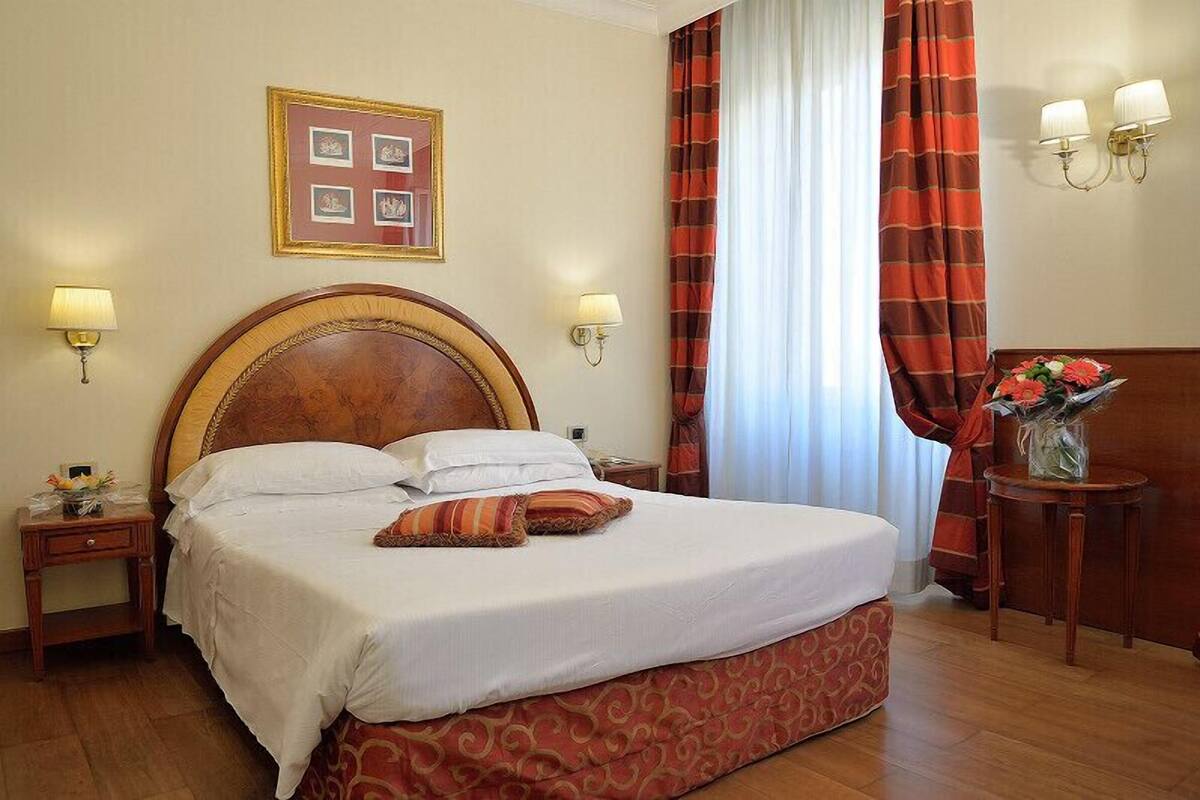 Una camera d'albergo con un letto grande, due abat-jour, un quadro appeso alla parete e una finestra con tende.