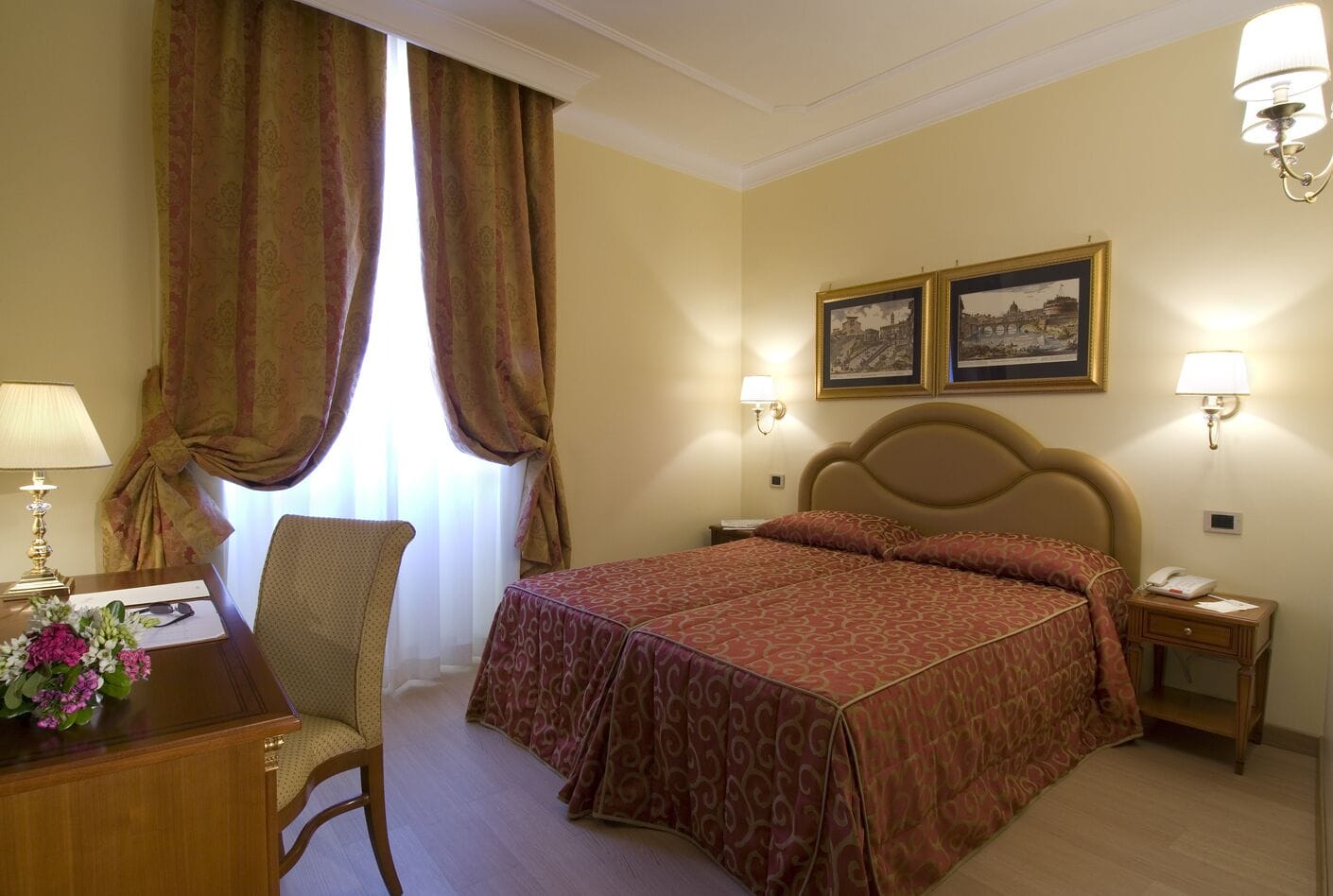 A hotel room with a large bed, a desk with a lamp, a chair, and two framed pictures on the wall.