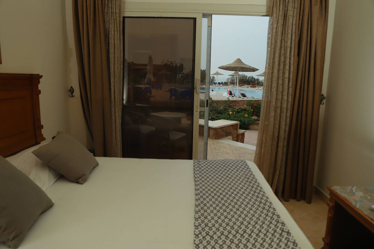 A hotel room with a bed, a patterned throw, a wooden nightstand, and a sliding glass door leading to a pool area.