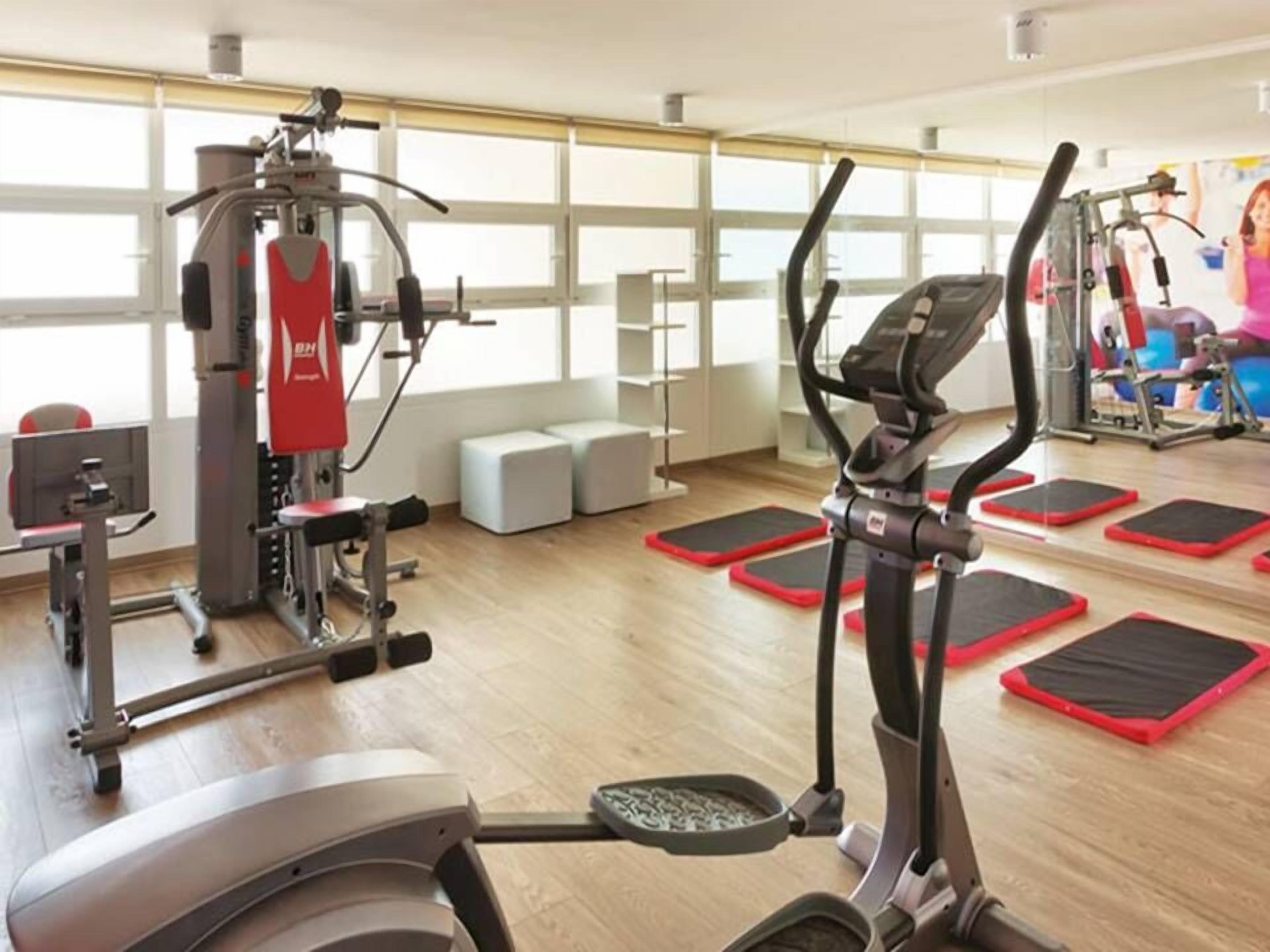 Un gimnasio con equipo de cardio, máquinas de pesas y colchonetas rojas en el suelo.