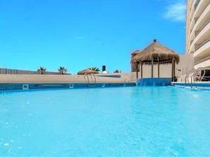 A clear blue pool with a thatched-roof structure, palm trees, and a modern building in the background. - Departamento con Piscina y Spa en la Serena (La Serena)