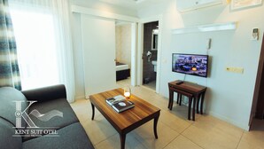Una habitación de hotel moderna con sofá, una mesa de centro de madera con libros, televisión y baño visible a través de una puerta abierta.