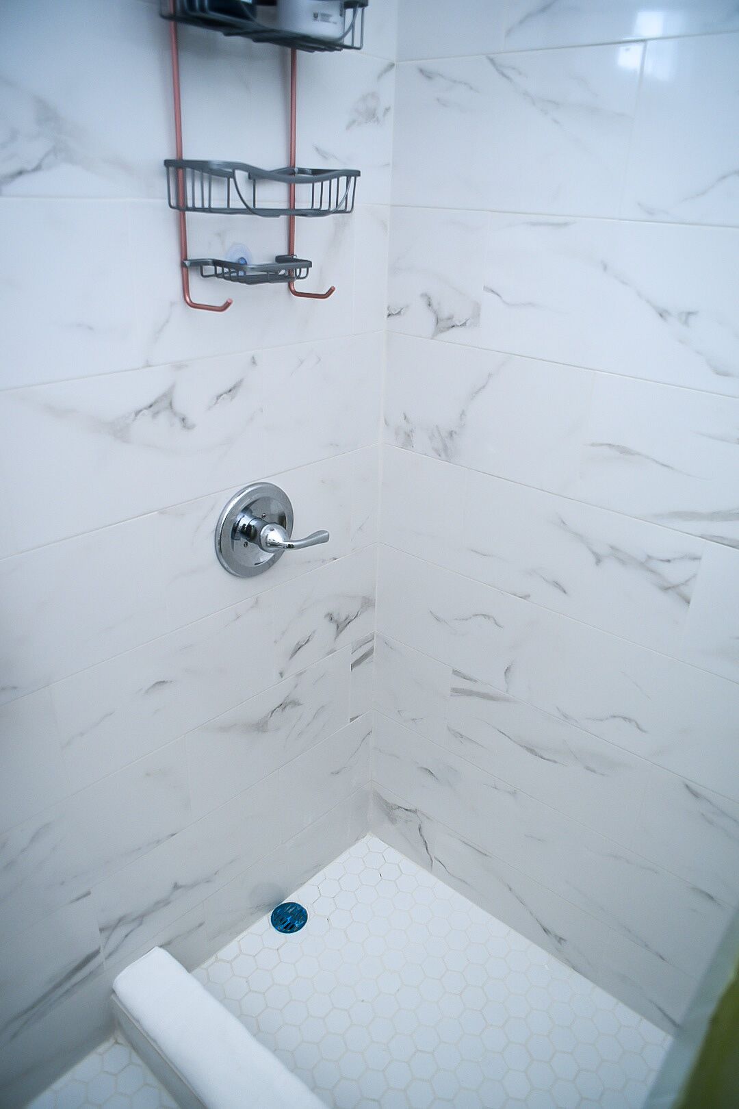 Un baño con baldosas de mármol blanco, una ducha con grifo de cromo y un pequeño estante con artículos de aseo.