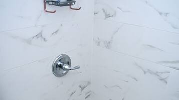Un baño con baldosas de mármol blanco, una ducha con grifo de cromo y un pequeño estante con artículos de aseo.