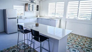 A compact kitchen with a central island, two bar stools, a refrigerator, and a window with blinds. - La Ferreteria Apartments (San Juan)