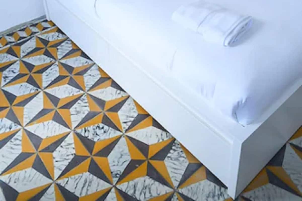 Un lit bien fait, avec du linge de lit blanc et une serviette enroulée, une lampe de chevet et un sol orné d’un motif géométrique.