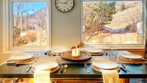 Una mesa de comedor preparada para cuatro personas, con un reloj en la pared y vistas a un paisaje montañoso a través de dos ventanas.