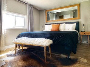Un dormitorio con una cama grande, un banco de madera, una mesita de noche, un espejo y una ventana con cortinas.