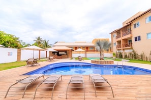 A swimming pool with lounge chairs, a bar area, and a building with balconies. - GB37 Excelente Apt 2 Suítes em Guarajuba (Camaçari)