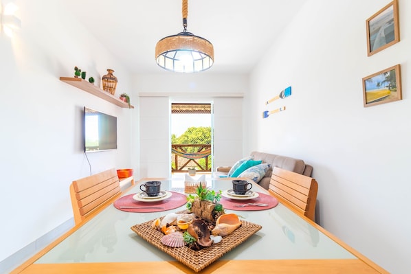 A dining area with a glass table, wicker placemats, and a central decorative piece. There are two wooden chairs, a sofa, and a television mounted on the wall. - GB37 Excelente Apt 2 Suítes em Guarajuba (Camaçari)
