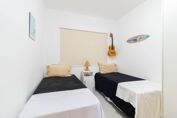 Ein kleiner, sauberer Raum mit zwei Einzelbetten, einem Nachttisch mit Lampe, einer Gitarre und einem Surfbrett an der Wand.