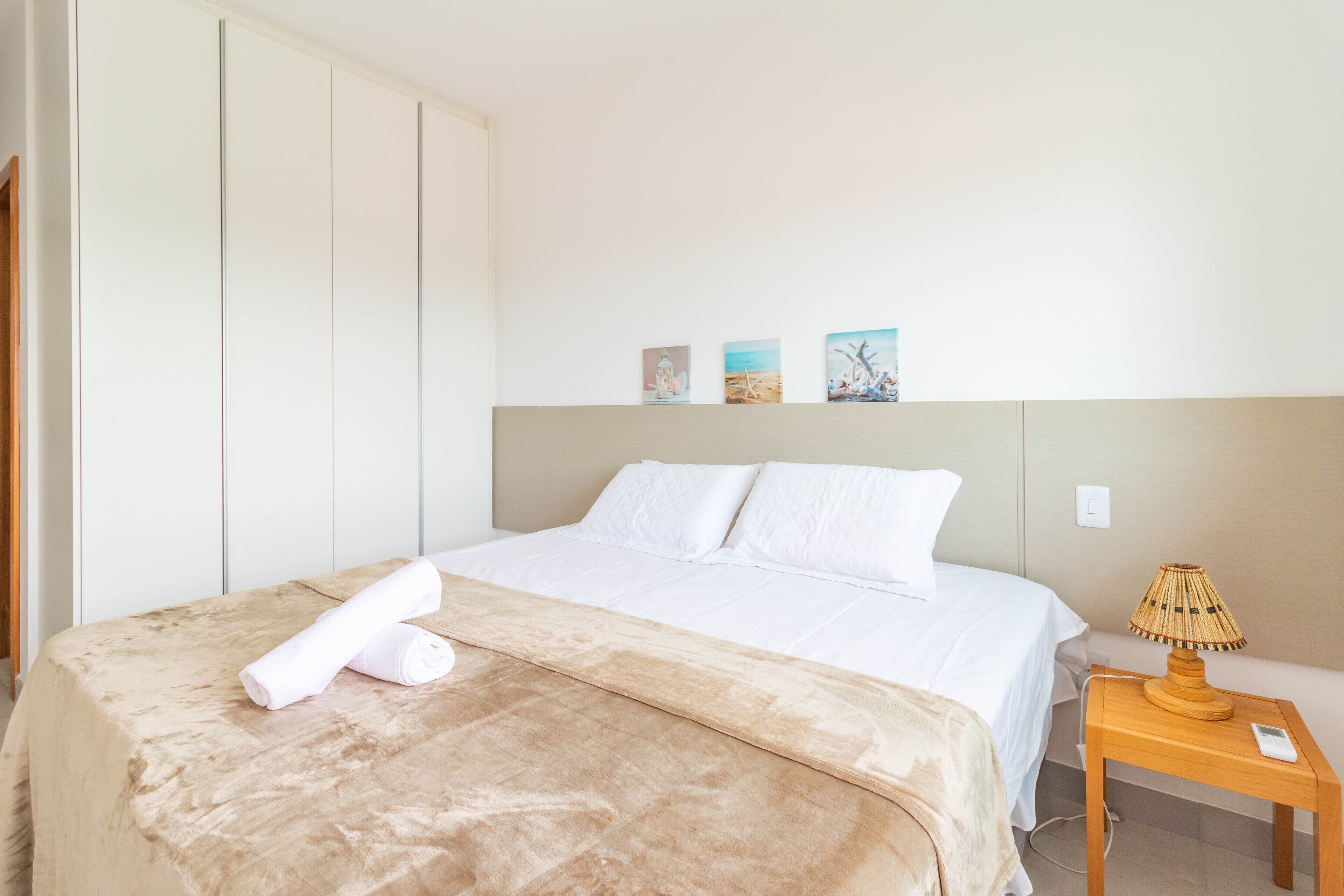 Ein ordentlich bezogenes Bett mit einer beigen Decke, ein Nachttisch aus Holz mit einer Lampe und drei kleine Bilder an der Wand.