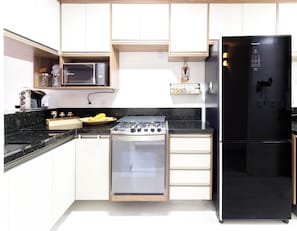 A modern kitchen with white cabinets, a black refrigerator, a microwave, a gas stove, and a built-in oven. - GB37 Excelente Apt 2 Suítes em Guarajuba (Camaçari)