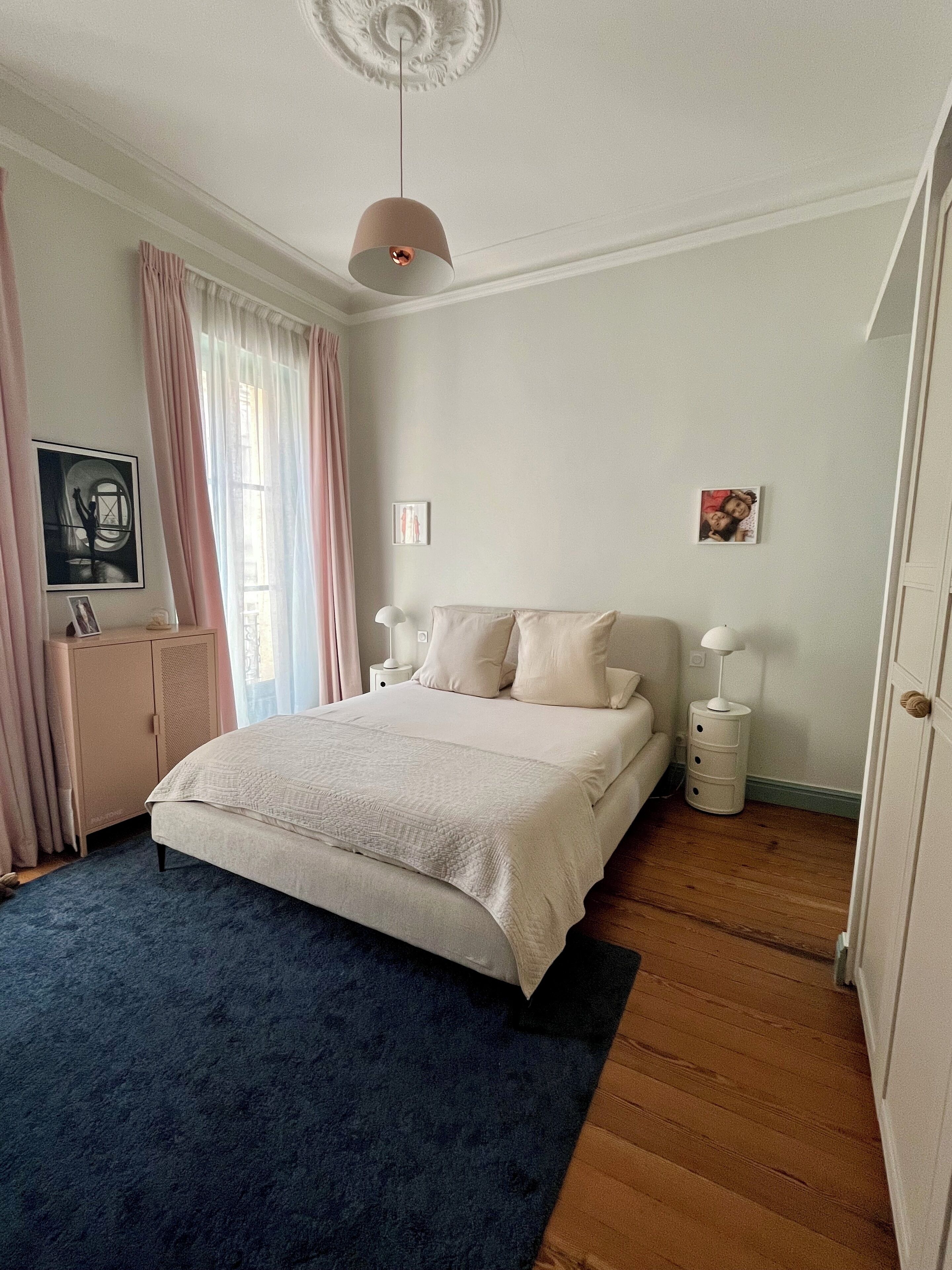 Une chambre à coucher comprenant un lit, des tables de chevet, une armoire, un luminaire au plafond et une fenêtre avec des rideaux.