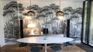 Ein Essbereich mit einem runden Tisch, Stühlen aus Drahtgeflecht, einer weißen Tischlampe und einem Wandbild von Bäumen.