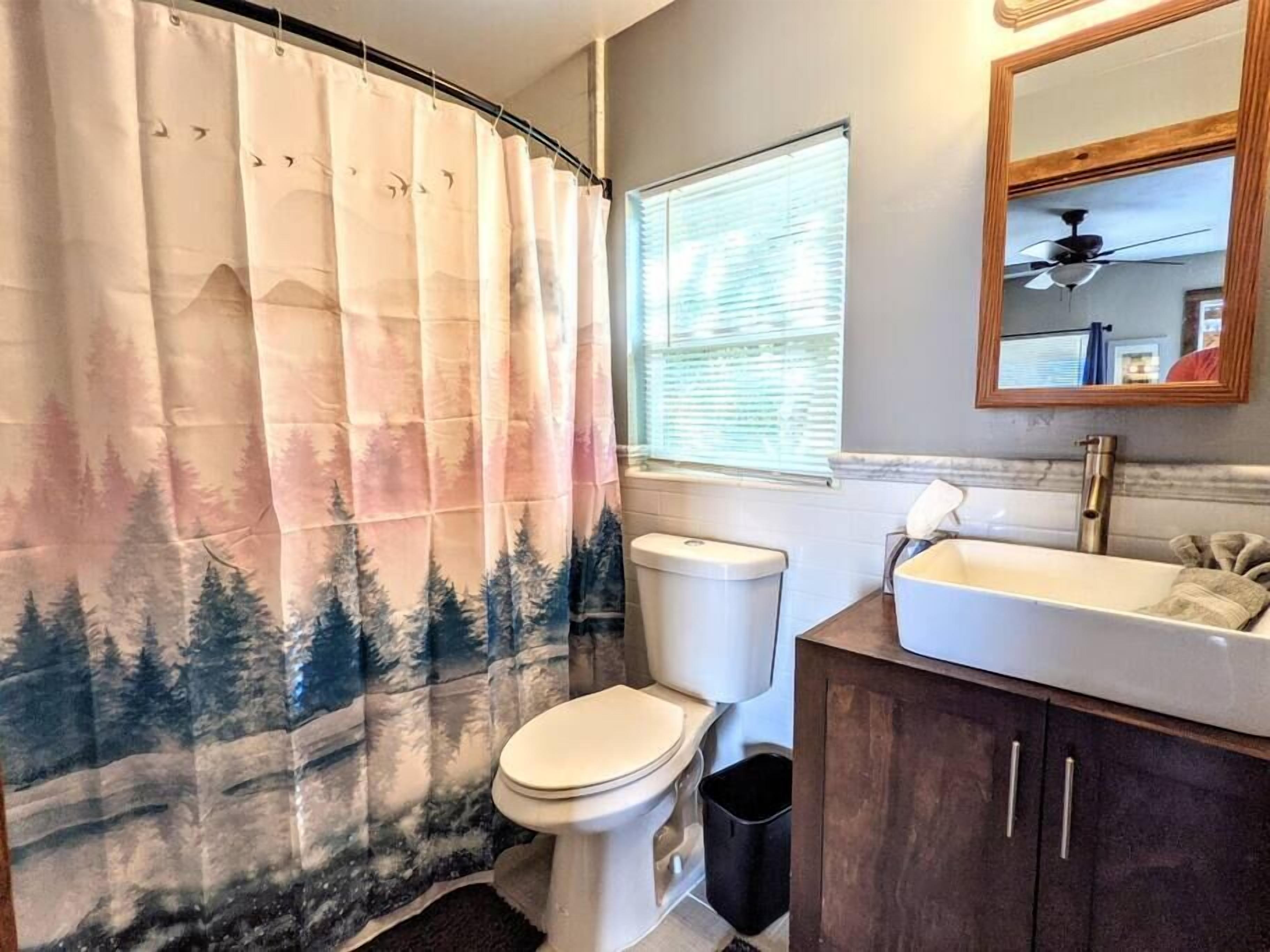 Un baño con cortina de ducha temática de bosque, un inodoro, un lavamanos y una ventana con persianas.