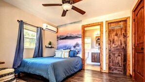 Ein Schlafzimmer mit Bett, Deckenventilator, Klimaanlage, Holzboden und einer Tür zu einem Badezimmer.