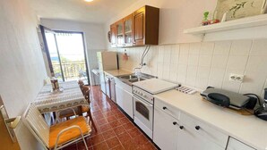 Een kleine keuken met witte kastjes, een fornuis en een spoelbak. Er staat een eettafel met stoelen en een uitzicht door een raam.